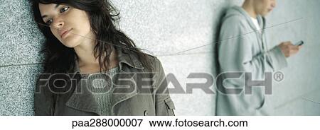 Stock Photo - Young woman leaning against wall while young man looks at cell phone. Fotosearch