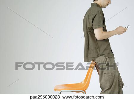 Young man leaning on back of chair, holding cell phone View Large Photo Image Stock Image - Young man leaning on back of chair, holding cell phone. Fotosearch