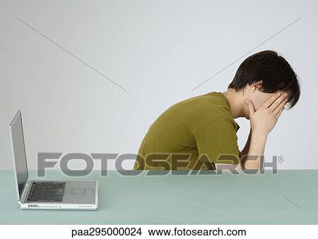 Young man sitting next to laptop, holding head in hands View Large Photo Image Picture - Young man sitting next to laptop, holding head in hands. Fotosearch