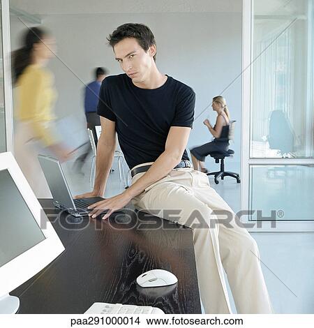 Man sitting on edge of desk in busy office View Large Photo Image Picture - Man sitting on edge of desk in busy office. Fotosearch