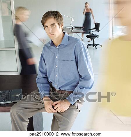 Stock Image - Man sitting on edge of desk in busy office. Fotosearch