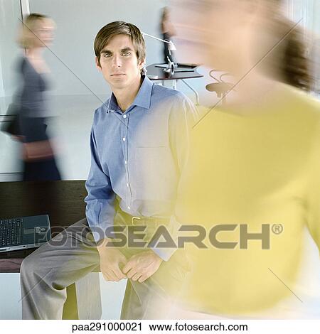 Stock Image - Man sitting on edge of desk in busy office. Fotosearch