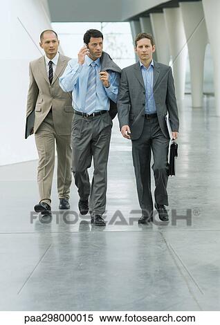 Three businessmen walking through lobby, one using cell phone View Large Photo Image Stock Photography - Three businessmen walking through lobby, one using cell phone. Fotosearch