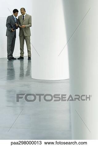 Stock Image - Two businessmen standing in lobby, looking at cell phone. Fotosearch