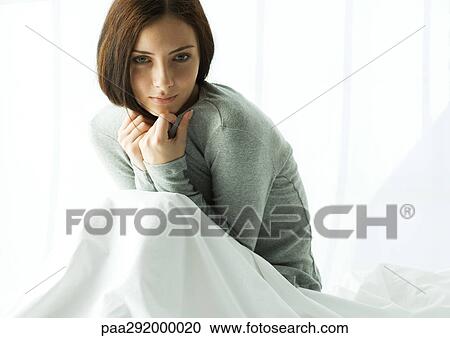 Stock Image - Young woman sitting up in bed, curled up. Fotosearch