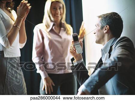 Businessman holding up cellphone, female colleagues smiling and clapping View Large Photo Image Picture - Businessman holding up cellphone, female colleagues smiling and clapping. Fotosearch