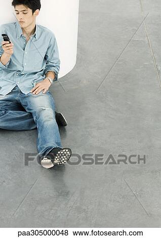 Young man sitting on floor looking at cell phone View Large Photo Image Stock Photo - Young man sitting on floor looking at cell phone. Fotosearch