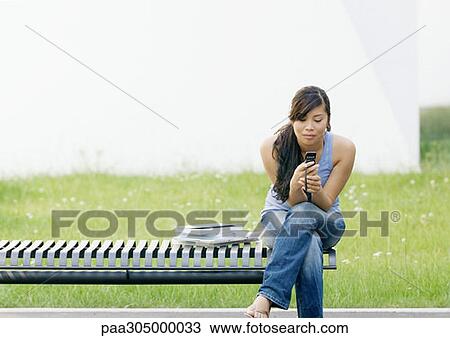 Young woman sitting on end of bench looking at cell phone View Large Photo Image Stock Image - Young woman sitting on end of bench looking at cell phone. Fotosearch