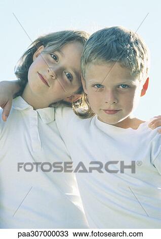 Stock Image - Boy and girl standing with arms around each other's shoulders, portrait. Fotosearch