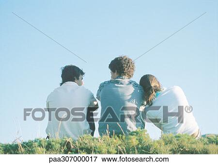 Stock Photo - Three teenagers sitting on grass, girl leaning head on boy's shoulder. Fotosearch