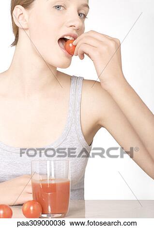 Stock Photograph - Young woman eating tomato, with glass of tomato juice. Fotosearch
