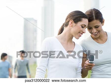 Two young women looking down at cell phone in urban park View Large Photo Image Stock Image - Two young women looking down at cell phone in urban park. Fotosearch