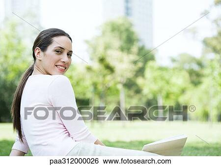 Stock Photograph - Young woman sitting on grass with book in urban park. Fotosearch