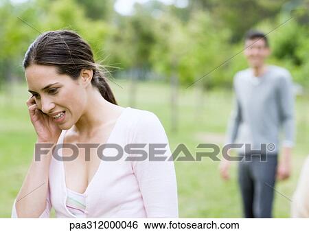 Young woman talking on cell phone, man in background, in park View Large Photo Image Stock Photograph - Young woman talking on cell phone, man in background, in park. Fotosearch