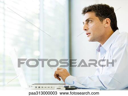 Stock Image - Man sitting at laptop, looking out window. Fotosearch