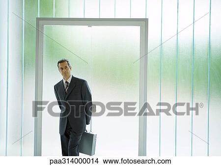 Picture - Businessman standing in front of door, holding briefcase behind back. Fotosearch