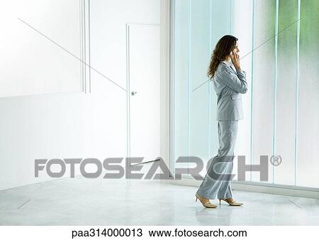 Stock Image - Businesswoman standing near window, using cell phone. Fotosearch