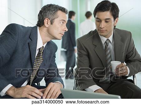 Stock Image - Two businessmen sitting around laptop, one with coffee cup. Fotosearch