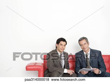 Stock Photo - Two businessmen sitting on couch, one holding document. Fotosearch