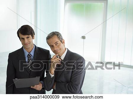 Two businessmen standing side by side, one holding file View Large Photo Image Stock Photo - Two businessmen standing side by side, one holding file. Fotosearch