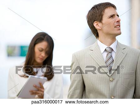 Stock Photo - Businessman looking up while female assistant takes notes in background. Fotosearch