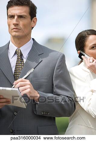 Businessman taking notes while businesswoman uses cell phone View Large Photo Image Picture - Businessman taking notes while businesswoman uses cell phone. Fotosearch
