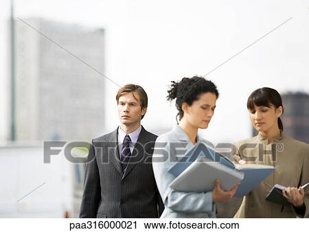 Stock Image - Two businesswomen discussing file, businessman passing by in background. Fotosearch