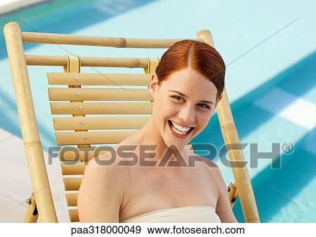 Stock Photo - Woman sitting in deckchair smiling, pool in background. Fotosearch
