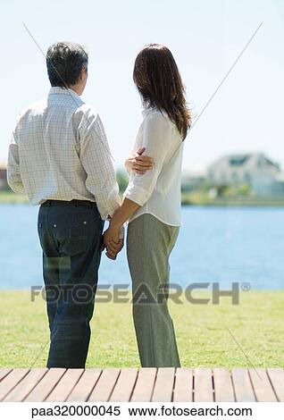 Stock Photography - Couple standing and looking at lake, holding hands. Fotosearch