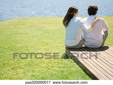 Stock Photo - Mature couple sitting at end of wooden walkway, near water. Fotosearch