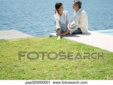 Stock Image - Mid-adult couple sitting by edge of water. Fotosearch