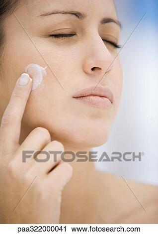 Stock Image - Woman applying moisturizer to cheek, eyes closed. Fotosearch