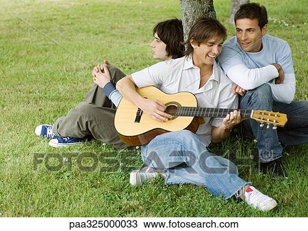 Young male friends sitting near tree, playing guitar View Large Photo Image Stock Image - Young male friends sitting near tree, playing guitar. Fotosearch