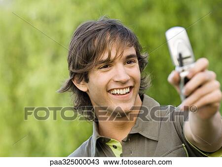 Stock Image - Young man holding up cell phone and smiling. Fotosearch