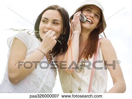 Two young woman outdoors, one using cell phone View Large Photo Image Stock Photo - Two young woman outdoors, one using cell phone. Fotosearch