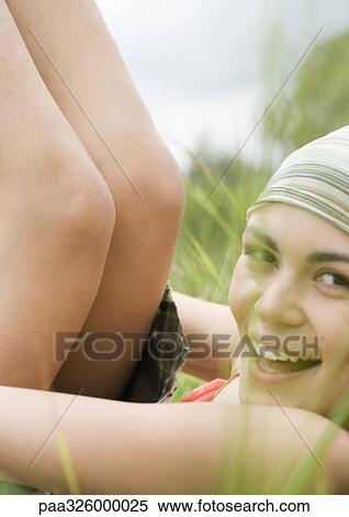 Stock Photography - Young woman lying in grass with legs up, smiling at camera. Fotosearch
