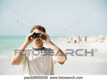 Stock Image - Man looking through binoculars on beach, facing camera. Fotosearch