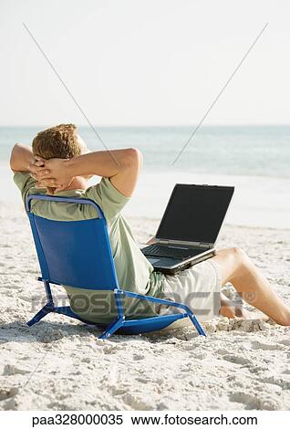 Stock Photography - Man sitting on beach with computer on lap. Fotosearch