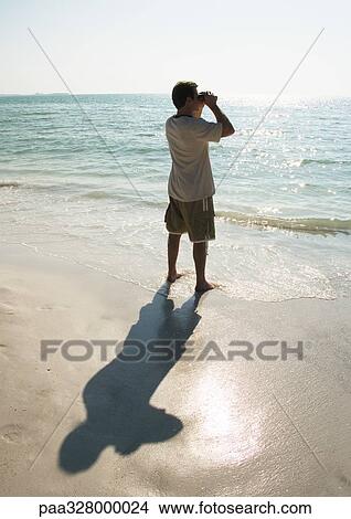 Man standing on beach, looking out to sea with binoculars View Large Photo Image Picture - Man standing on beach, looking out to sea with binoculars. Fotosearch