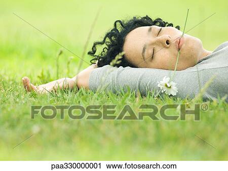 Stock Image - Young woman lying on grass with flower in mouth and eyes closed. Fotosearch