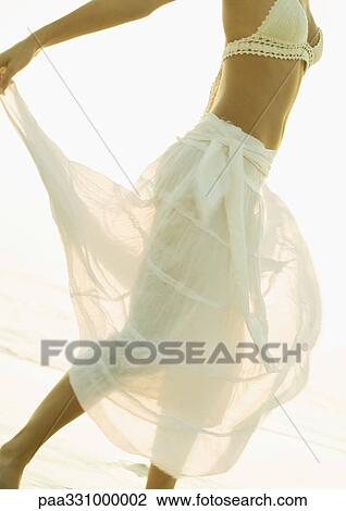 Stock Image - Woman twirling translucent skirt on beach, chest down. Fotosearch