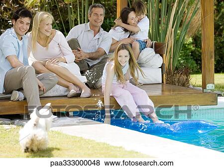 Stock Photo - Extended family sitting by edge of pool, girl splashing water at dog. Fotosearch