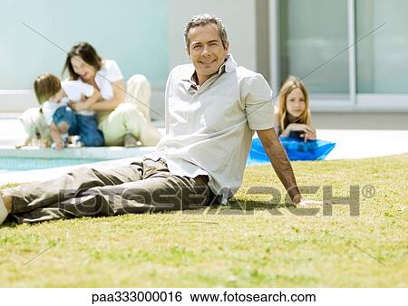 Stock Photograph - Man relaxing on grass, family in background. Fotosearch