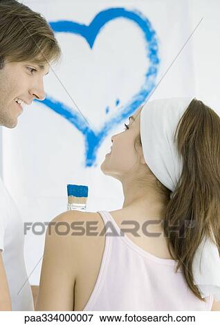 Stock Photo - Couple smiling at each other, heart painted on wall in background. Fotosearch