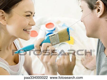 Stock Image - Woman holding up paintbrush toward man's face. Fotosearch