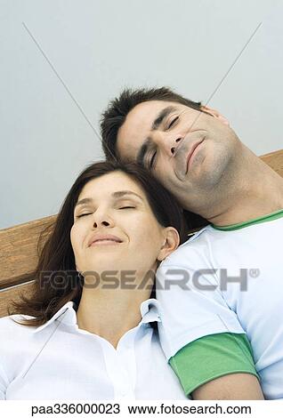 Stock Image - Couple resting on a bench, reclining with eyes closed and heads together, smiling, portrait. Fotosearch
