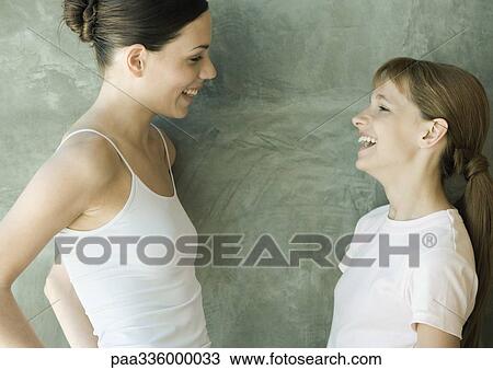 Stock Image - Two sisters standing, looking at each other and laughing, side view. Fotosearch