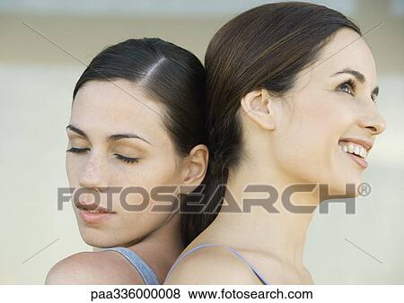 Stock Photo - Two young women back to back, one with closed eyes while the other one is smiling, close-up. Fotosearch