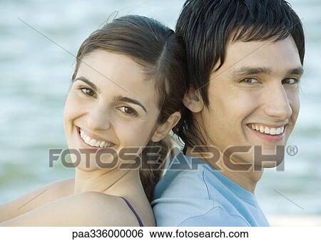 Stock Photograph - Young couple sitting back to back near water, both smiling. Fotosearch