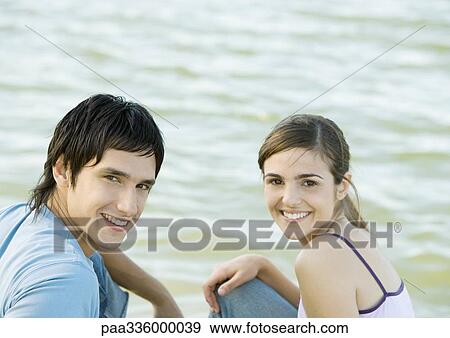 Young couple sitting side by body of water, smiling and looking at camera View Large Photo Image Stock Photo - Young couple sitting side by body of water, smiling and looking at camera. Fotosearch